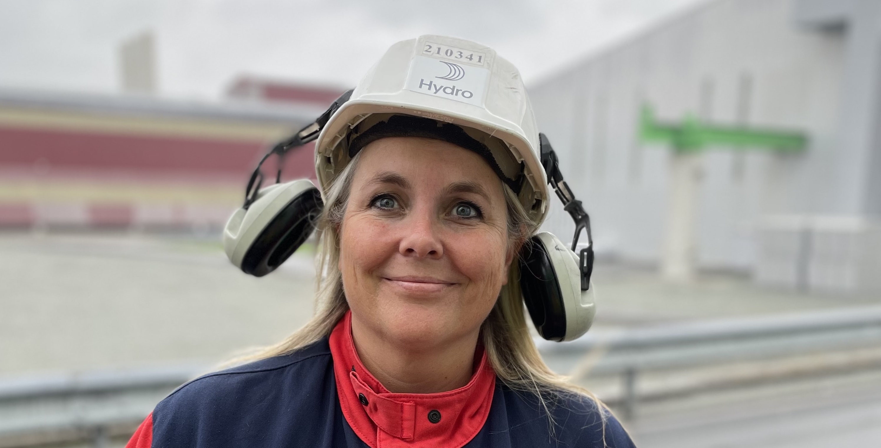 Kvinne med industriarbeidsklær og hjelm smiler til kamera. Foto.