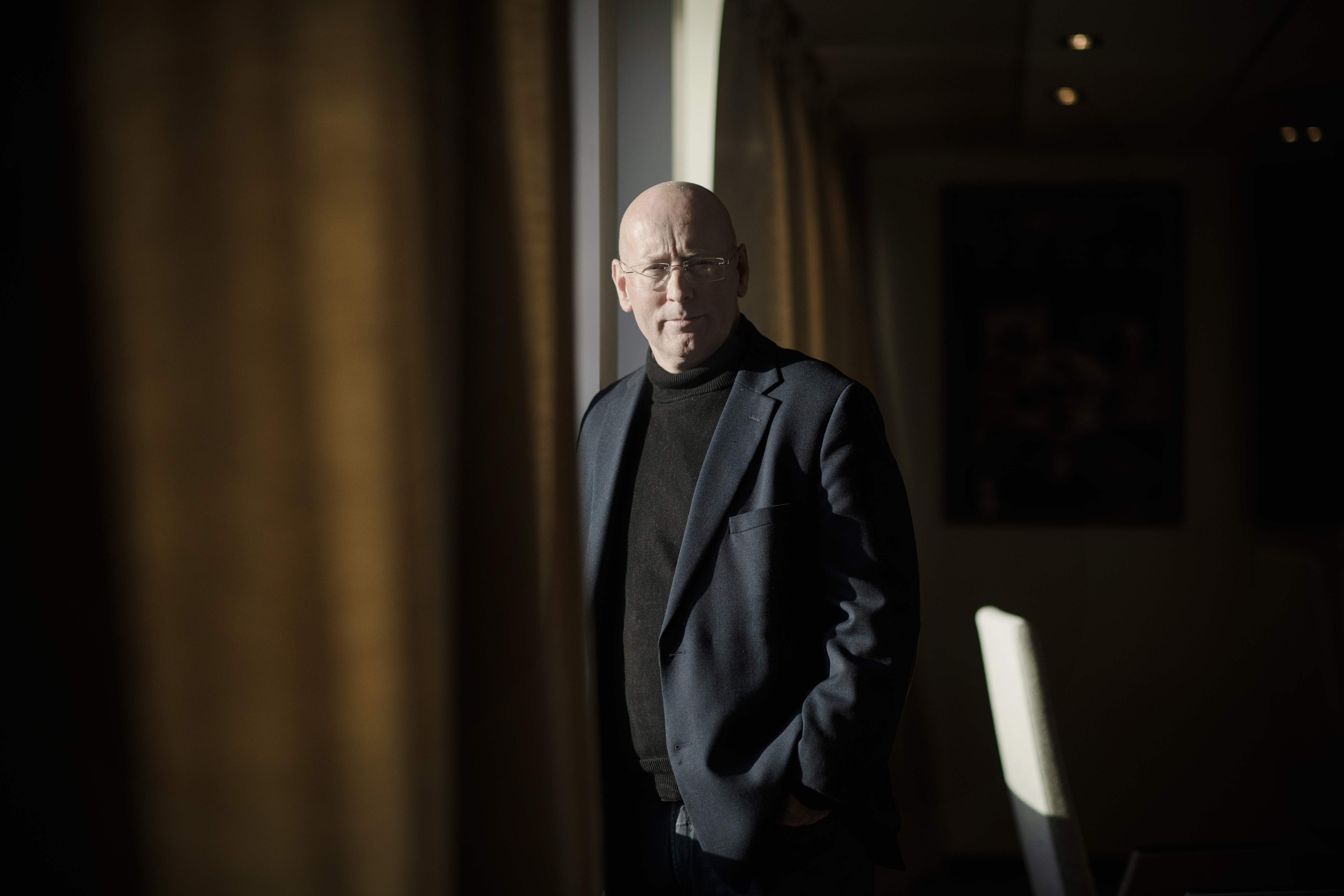 Mann i dress står ved vindu, mellom gardiner og ser mot kamera. Foto